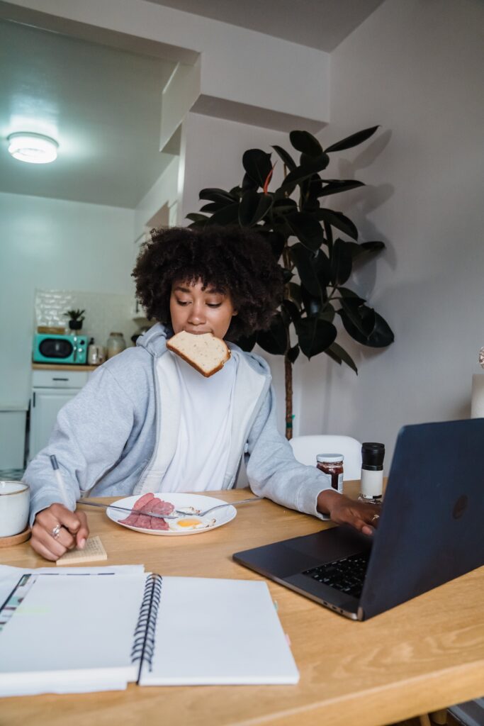 A women sitting at a table juggling eating, working on a computer, and using a computer.