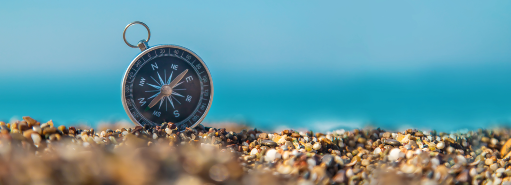 Close-up of a weathered compass resting on the sandy shore of a beach, with grains of sand partially covering its base. The compass needle points north, and the surrounding scenery hints at a peaceful, natural setting by the ocean.