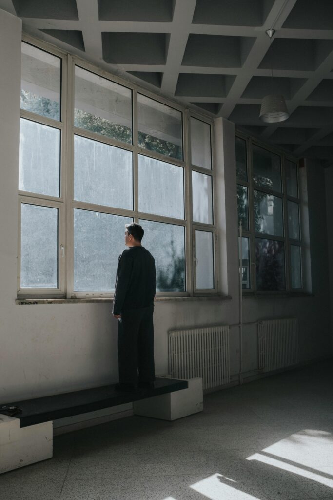 A person stands alone in a dimly lit room, gazing out of a large window with a blank, contemplative expression. The room has an industrial, minimalist look, with pale walls, tiled flooring, and a radiator beneath the window. The atmosphere feels introspective and somber, as the individual appears deep in thought, surrounded by silence.