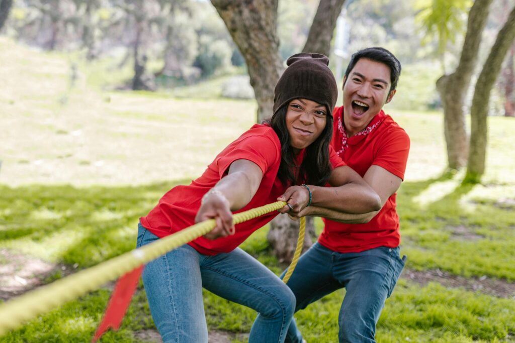 Two people engaged in a tug-of-war, both wearing red shirts, with determined expressions on their faces as they pull on a yellow rope in an outdoor setting.