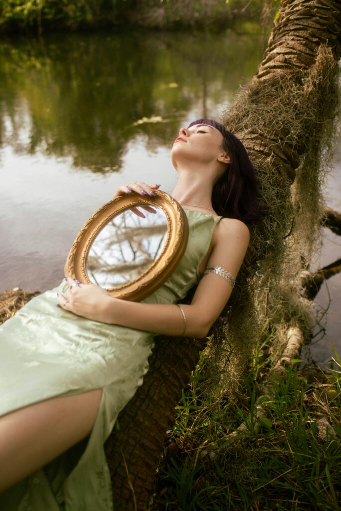 The image shows a woman reclining against a tree by a calm body of water, holding a round golden mirror in her lap. She is dressed in a light green satin gown and wears an arm cuff. Her head is tilted back with her eyes closed, appearing serene and reflective. The mirror in her hand reflects the trees and sky above, adding a dreamy, introspective quality to the scene. The natural, peaceful setting enhances the sense of calm and contemplation in the image.