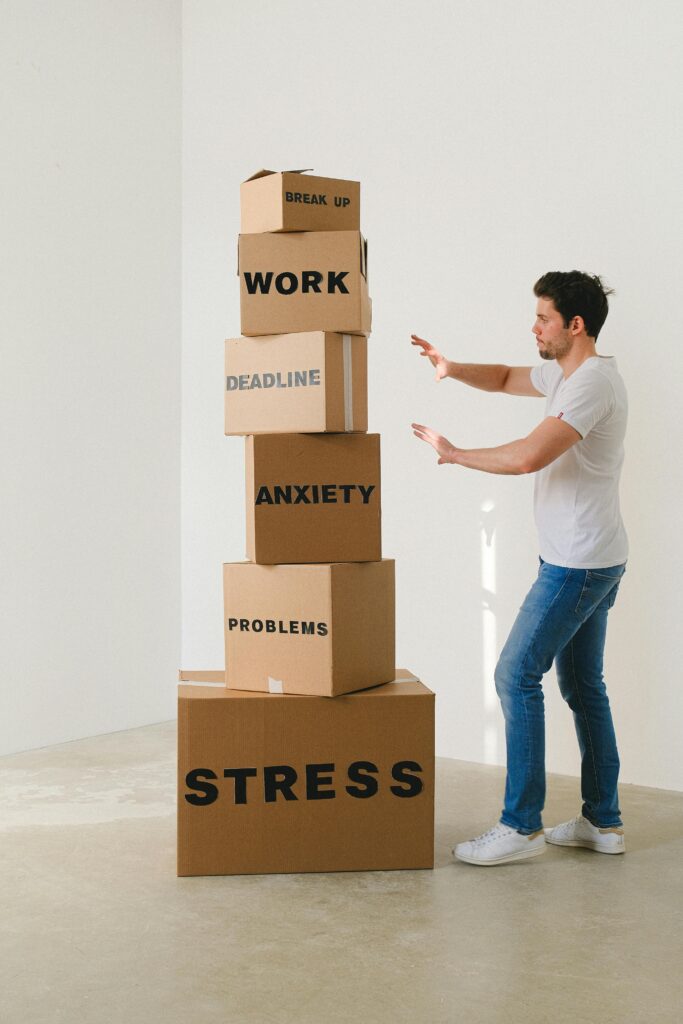 The image shows a man standing beside a tall stack of cardboard boxes arranged one on top of the other. Each box has a word printed on it, indicating various stressors. From bottom to top, the boxes are labeled 